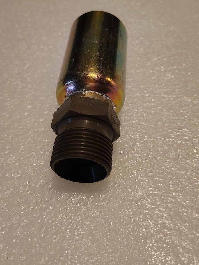 A Weatherhead 12U-P62 hydraulic fitting, a metallic cylindrical valve with a threaded end and a shiny, rainbow-like finish, is resting on a textured white surface.