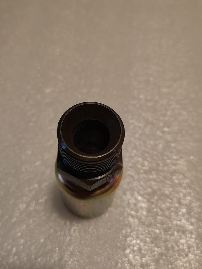 A metallic cylindrical piece with a threaded top lies on a white textured surface, its reflective sheen suggesting it is the Weatherhead 12U-P62, a 3/4 hose by 3/4 BSPT male fitting, known for use as a hydraulic hose fitting or connector.
