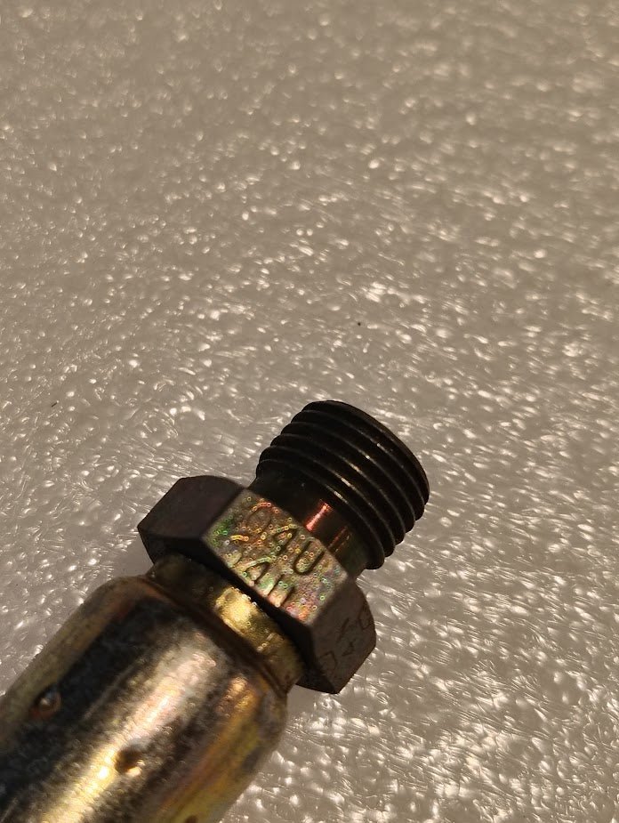 Close-up of the 04U-P54 Weatherhead fitting: a metallic hexagonal bolt with a threaded end on a textured surface. Its engraved text indicates its compatibility with Weatherhead fittings, enhancing the intrigue of its robust design.