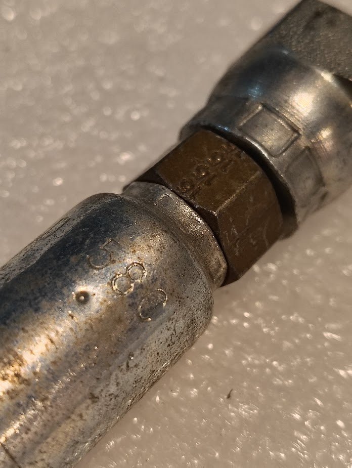 A close-up of a slightly worn Weatherhead 04U-58C connector with threaded ends on a textured surface, reminiscent of Parker hydraulic craftsmanship. Numbers and markings are engraved on its 1/4 Hose x M16x1.5 DIN Female Fitting - Straight Swivel, focusing on the foreground details.