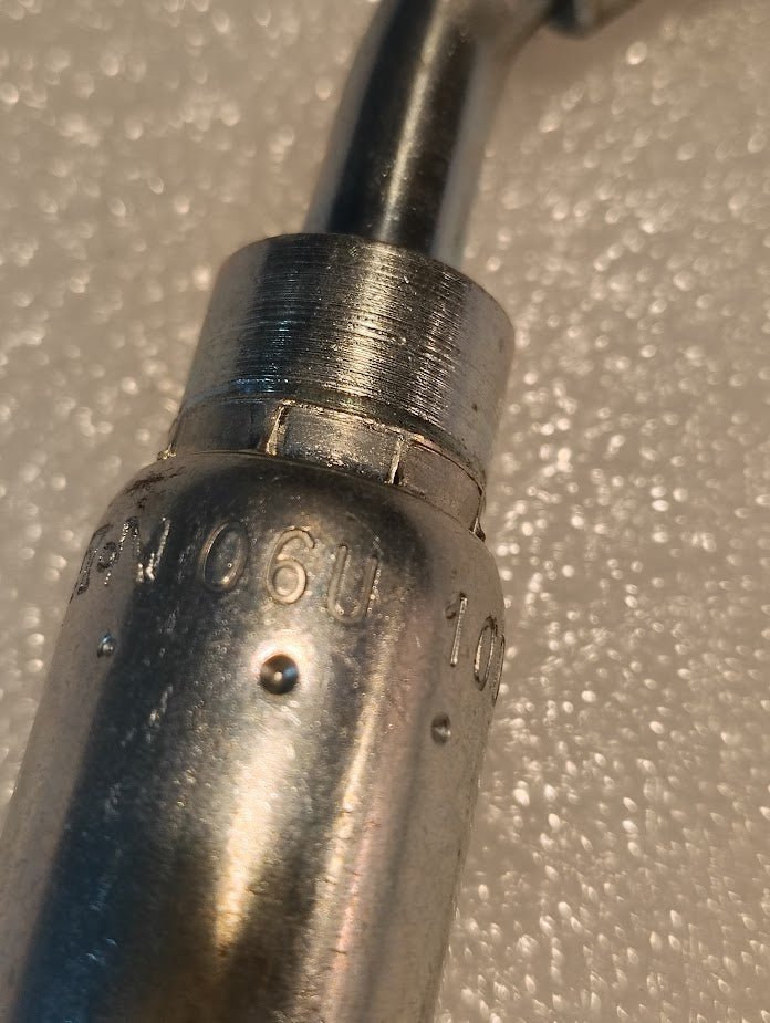 Close-up of a metallic 06U-10D Weatherhead fitting, engraved SN 06U 10, with a shiny surface contrasting against a textured white background, highlighting its use as an interchangeable component in advanced hydraulic hose systems.