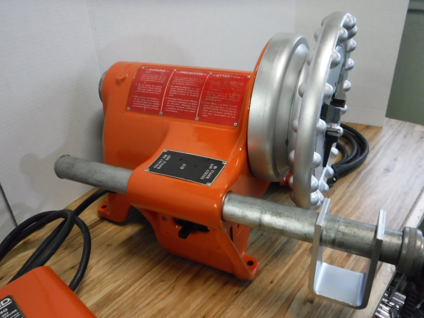 An Aeroquip FT1097-1-5, a reconditioned reusable hose assembly machine, is positioned on a wooden surface. This versatile orange tool comes equipped with a metal threading head, power cord, and multiple knobs and switches for precise operation, making it an excellent portable solution for industrial hose needs.