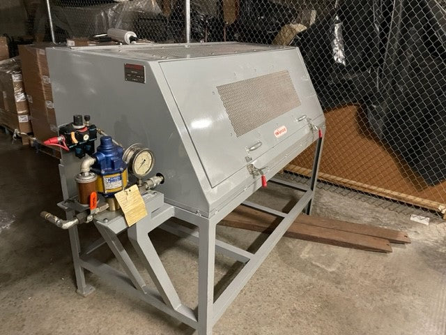 In the warehouse setting, a substantial machine stands out, closely resembling the Aeroquip FT1312 Hose Proof Test Stand. This used equipment boasts an air-driven power unit equipped with gauges and knobs on one side. The gray machine is characterized by its slanted top and mesh panel, set against a backdrop of chain-link fencing and cardboard boxes.