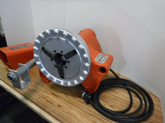 The Aeroquip FT1097-1-5 Reusable Hose Assembly Machine is a reconditioned industrial device in orange, featuring a circular metal component with multiple holes. It rests on a wooden surface, has a black cable extending from it, and is equipped with a sturdy metal handle on the side.