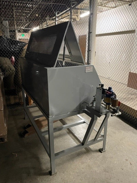 A large, industrial metal machine similar to an Aeroquip FT1312 Hose Proof Test Stand with a sloped top is positioned on a metal frame within a warehouse area enclosed by a chain-link fence. Pipes and valves are affixed to one side, while the dimly lit space contains various materials stored nearby.