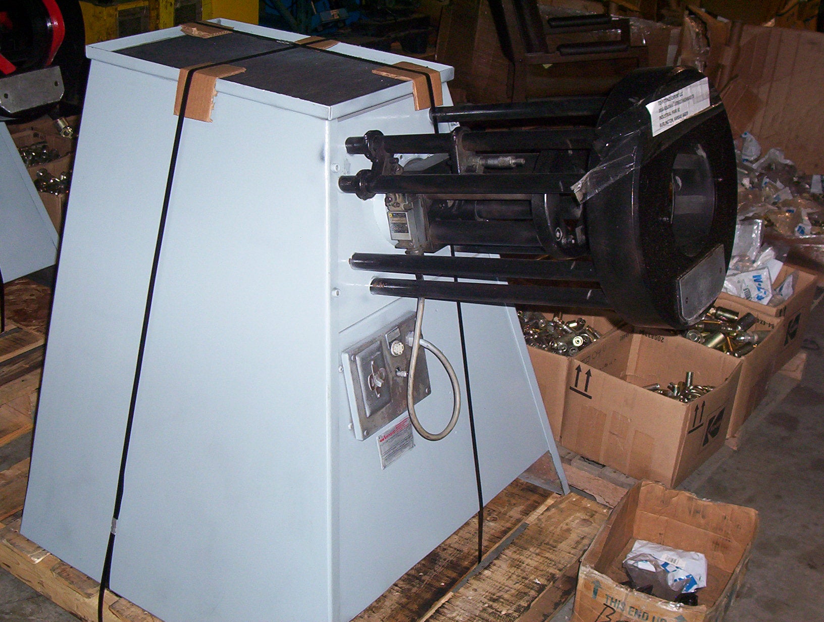 An Aeroquip FT1307 hydraulic 2" hose crimper, featuring a white, pyramid-shaped base and a rotating black mechanism on top, stands in the warehouse. Surrounded by cardboard boxes and various mechanical parts, this reconditioned machine with 10 dies is integral to efficient operations.