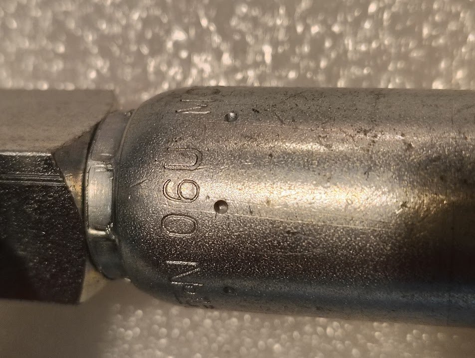 Close-up of a Weatherhead 06U-M06 (CS14-6X6) 3/8 Hose x 3/8 NPTF Male Fitting, a metallic cylindrical object with engraved markings, set against a textured white background.