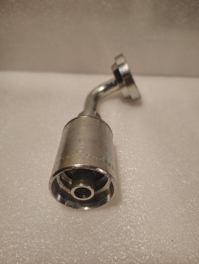 A close-up of the 43008U-G76 | 1/2 Hose x 1 Code 61 Flange Fitting - 90 bend (GW53-8x16), a shiny, metallic hydraulic fitting with a threaded end and curved section, placed on a white textured surface.