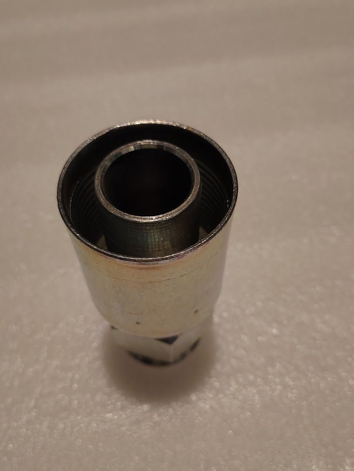 The 20U-520 Weatherhead fitting, a straight solid socket for a 1 1/4 hose with a 1 1/4 JIC male end, stands upright on a textured, light surface. Its cylindrical shape and threaded interior combine with its smooth and shiny exterior to softly reflect the light.