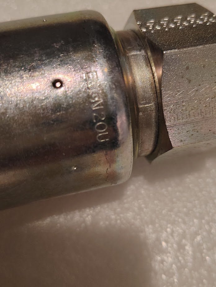 Close-up of a Weatherhead 20U-520 showing its shiny cylindrical design connected to a hexagonal nut. The surface has small dents and engraved letters, contrasted against the textured white background, highlighting its precision as a 1 1/4 Hose x 1 1/4 JIC Male Fitting - Straight Solid.
