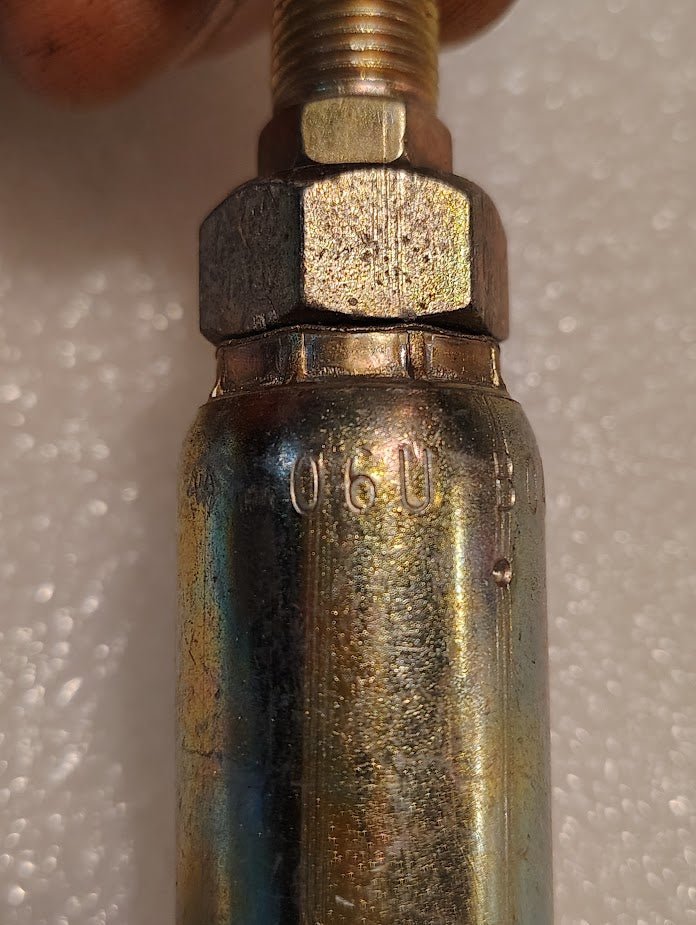 Close-up of a metallic cylinder marked 06U, part of the Weatherhead 06U-B04 series. The top has a threaded segment with a hexagonal nut for hydraulic hose fittings, against a textured white surface.