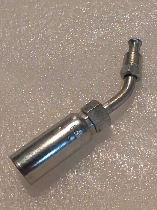 The 04U-B44 Weatherhead fitting, a small metallic component with a cylindrical body and bent, threaded end, lies on a textured white surface. It has a smooth, reflective finish and is designed for compatibility with 1/4 hydraulic hose fittings and 1/4 inverted male connections in a 45° swivel.