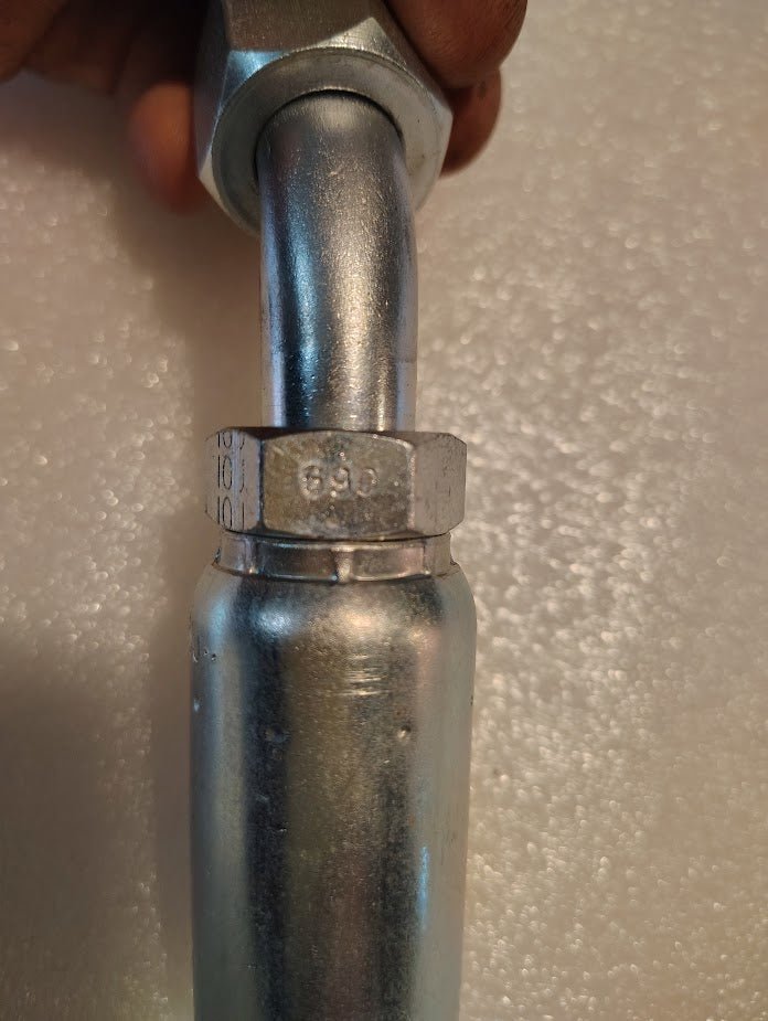 A close-up shows a shiny metal 10U-690 Weatherhead fitting, featuring a hex nut with engraved numbers. A hand grips the upper part against a textured light background.
