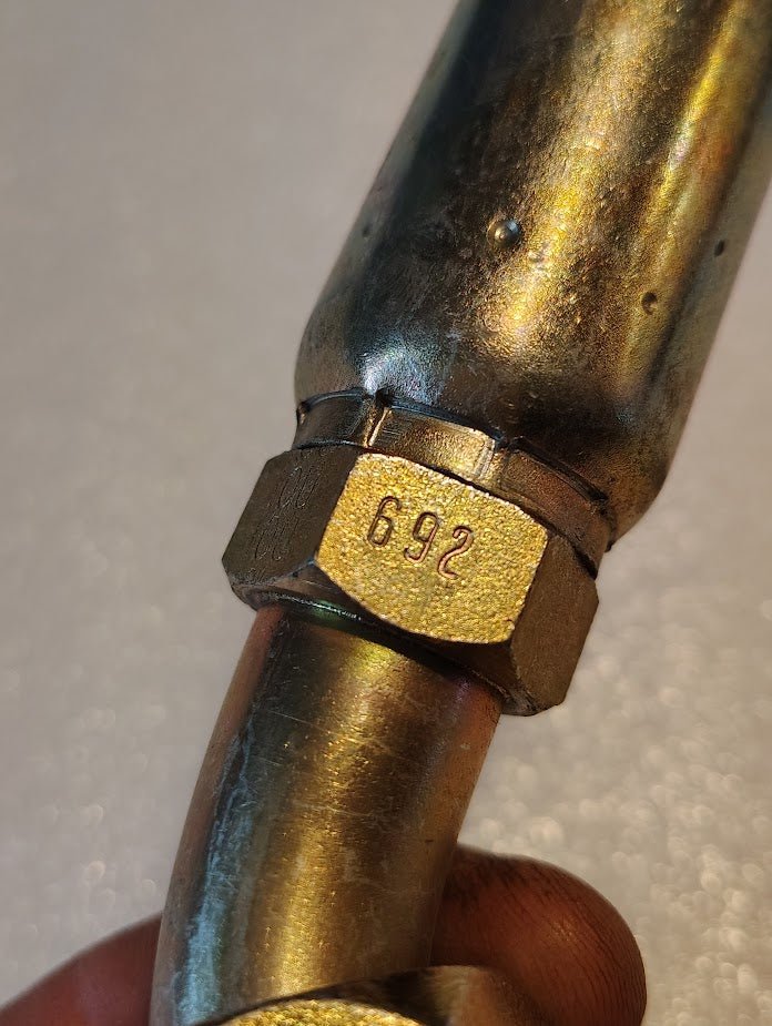 A close-up of a shiny 10U-692 metal fitting, compatible with Weatherhead fittings, shows a hexagonal nut with 692 engraved on it. The fitting is held by fingers against a blurred light background.