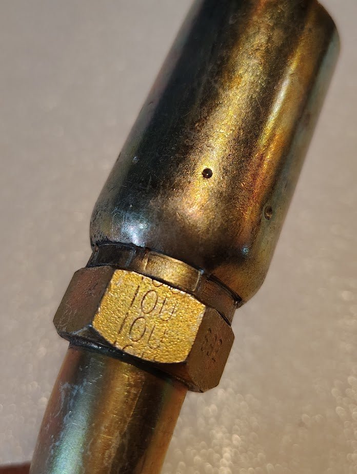 Close-up of a metallic component resembling a Weatherhead 10U-692 fitting, featuring a cylindrical body and hexagonal nut with engraved numbers. The surface appears slightly tarnished, while the background is softly blurred.