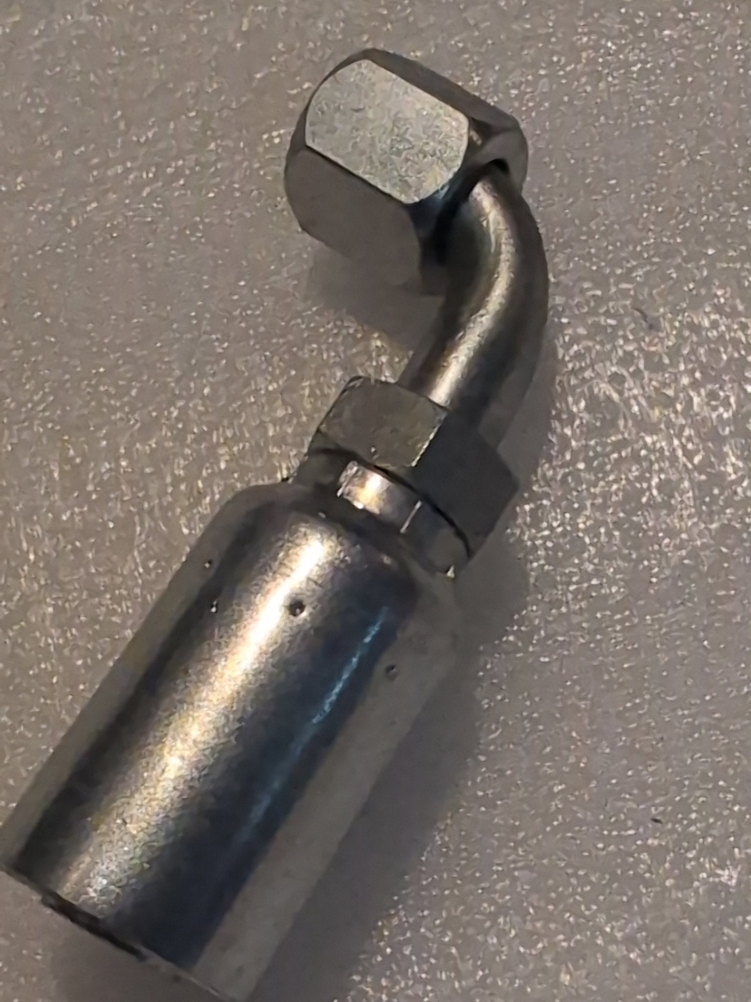 Close-up of a metallic Weatherhead 12U-670 fitting, compatible with 100R17 hoses, featuring a curved design and hexagonal nut, placed on a textured, light-colored surface.