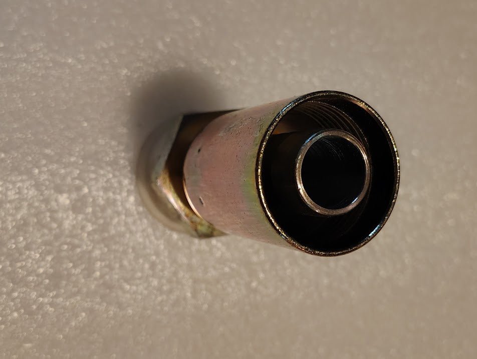Close-up of a metallic, cylindrical object resembling the Weatherhead 12U-E76 straight solid fitting, 3/4 hose x 1 ORF male. It has a dark interior and is mounted on a textured light surface with a slightly rusted exterior, suggesting industrial use.