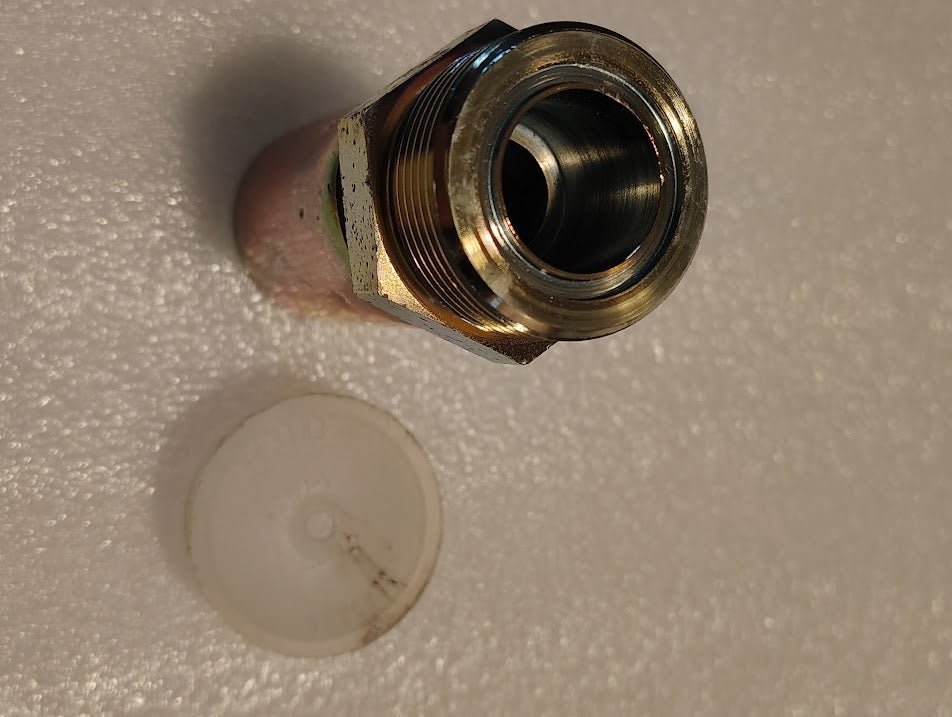 Close-up of a 12U-E76 Weatherhead 3/4 Hose x 1 ORF Male straight solid fitting on a textured white surface, beside a transparent plastic cap. The metal threaded hydraulic hose fitting appears unused, with light reflecting off its edges.
