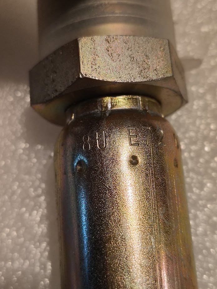 Close-up of a metallic connector, labeled 08U-E72, revealing its shiny surface. Resembling Weatherhead fittings, this 1/2 Hose x 3/4 ORF Male Fitting - Straight Solid stands out with an industrial aura against the textured white background.