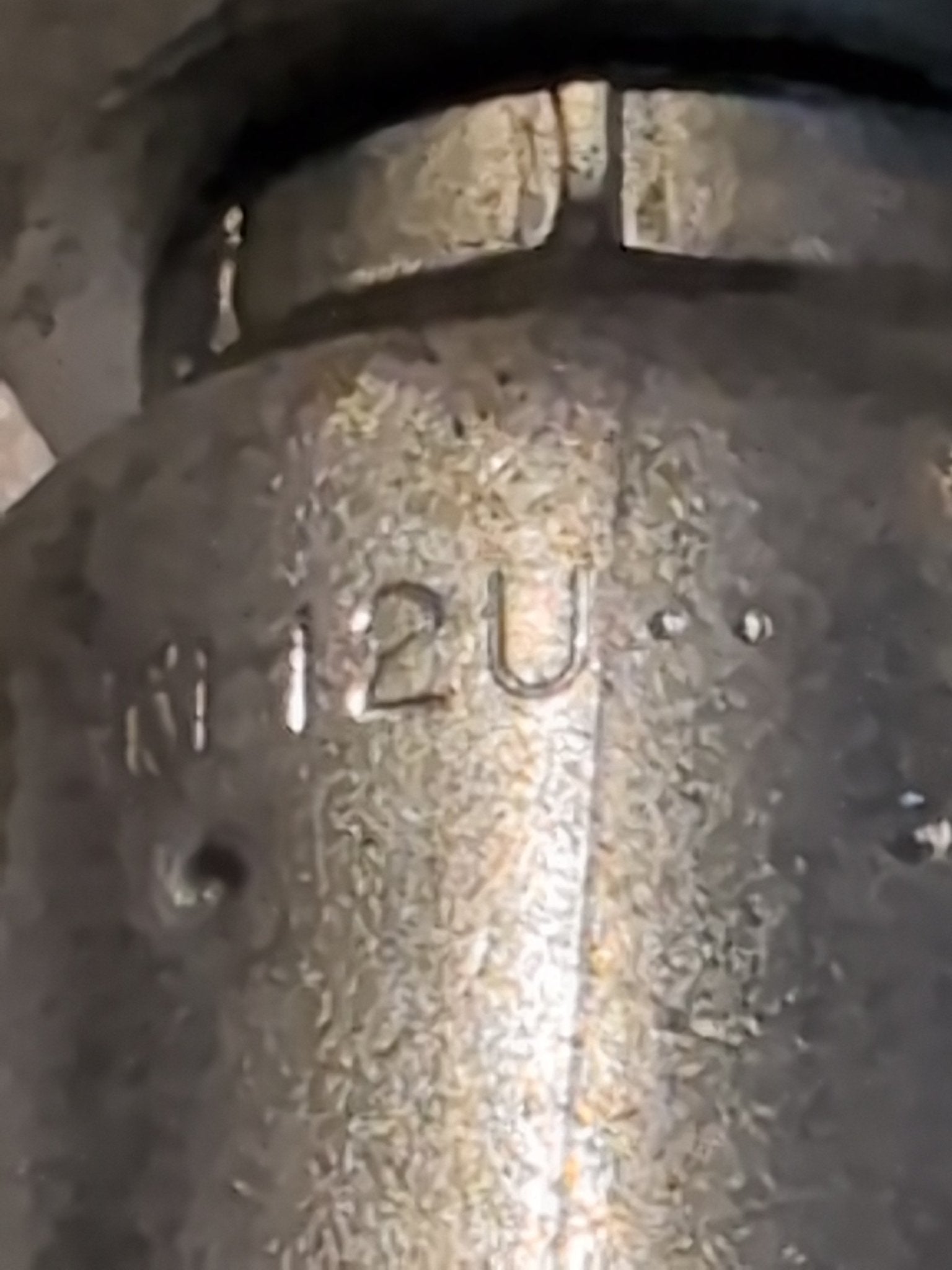 A close-up of a textured metal surface, similar to the Weatherhead 12U-S76 fitting, engraved with 12U and other indistinct marks. The slightly tarnished look suggests wear and age like that found on well-used hydraulic hose fittings.