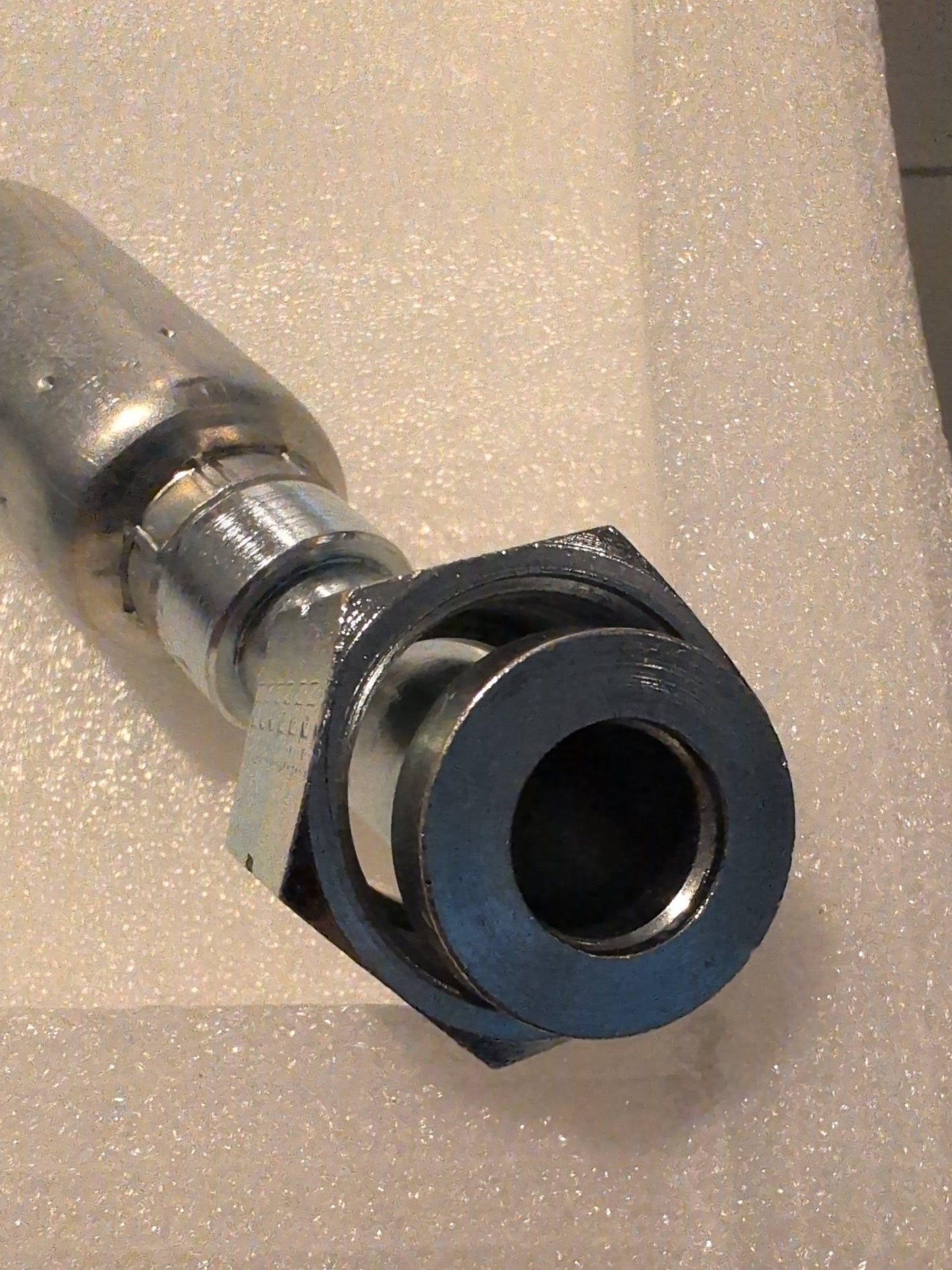 Close-up of a 12U-L72 Weatherhead fitting for 3/4 hose with a hexagonal nut on a textured white surface. The nut is shiny and reflective, while the background is softly blurred.