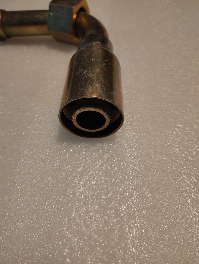 A 16U-A76 Weatherhead fitting, a 1 hose x 1 ORF female fitting with a rusty look, lies on a textured light surface. Its open circular end faces the viewer, featuring an angled joint to demonstrate its adaptability in various applications.