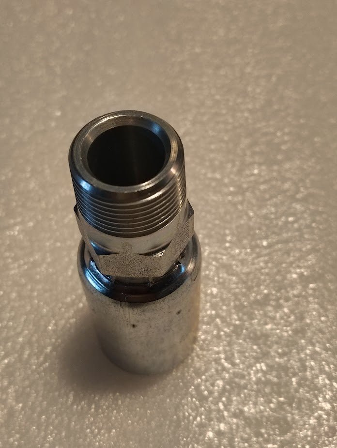 A shiny Weatherhead 12U-162 fitting, featuring 3/4 hose x 3/4 BSPT male connections, rests on a textured white surface. Its polished finish reflects light, exemplifying the high quality of Weatherhead fittings.