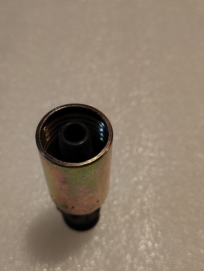 A close-up of a Weatherhead 06U-156 fitting, designed for 100R1 hoses, rests on a textured white surface. The cylindrical piece has a threaded interior and slightly tarnished exterior, with soft lighting casting shadows behind this essential metal plumbing component.