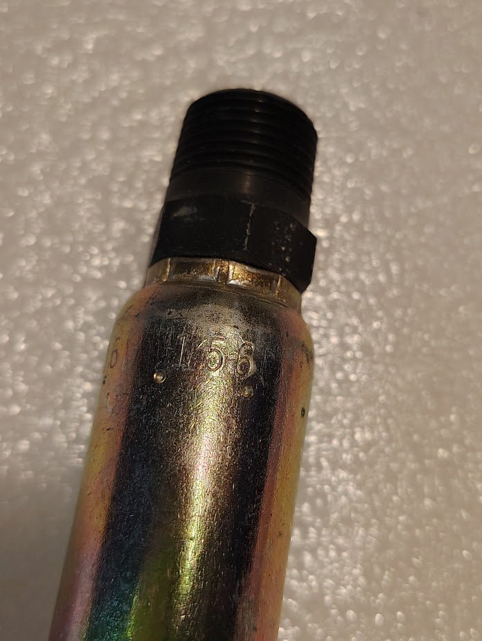 Close-up of a metallic cylindrical object with a threaded black end, probably a hydraulic hose fitting. The surface has engraved numbers, and the textured background displays small white circles, resembling Weatherheads 06U-156 fitting in industrial settings.