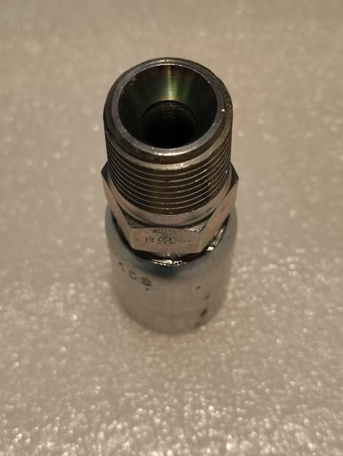 A close-up of the 08U-158 Weatherhead 1/2 Hose x 1/2 BSPT Male Fitting - Straight Solid, featuring metallic threads and a hollow center, rests on a textured, light-colored surface.