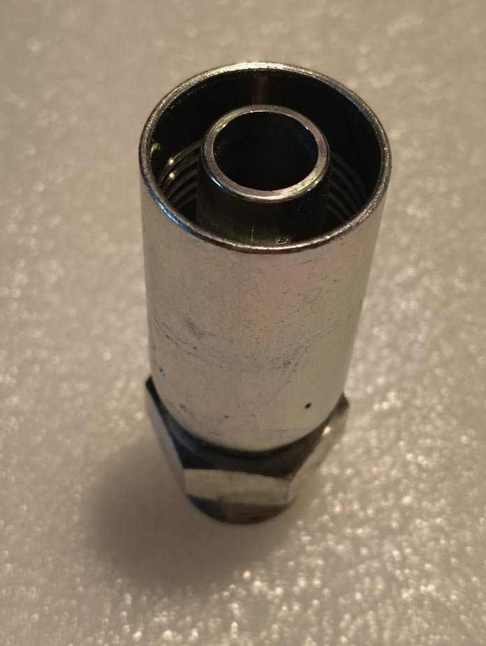 A close-up image of a metallic cylindrical object, the 10U-P60 Weatherhead 5/8 Hose x 5/8 BSPT Male Fitting - Straight Solid, is placed upright on a textured white surface. It features grooves inside the cylinder, indicating compatibility with interchangeable hydraulic hose fittings.