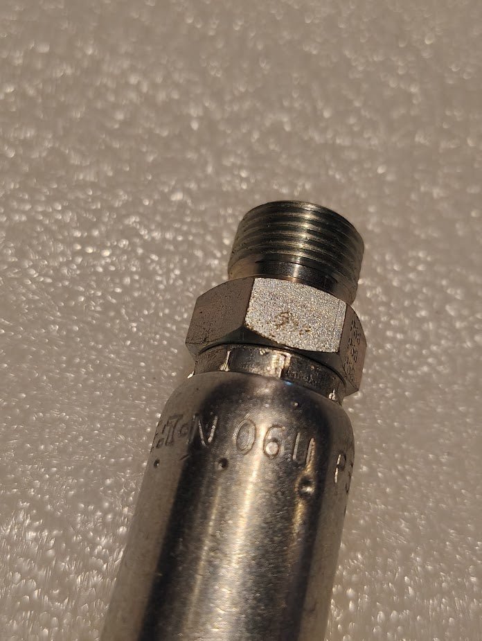 Close-up of the 06U-P56 | Weatherhead 3/8 Hose x 3/8 BSPT Male Fitting - Straight Solid, featuring a threaded end and engraved numbers. Resting on a textured light surface, lighting enhances its metallic finish and detailed engravings.
