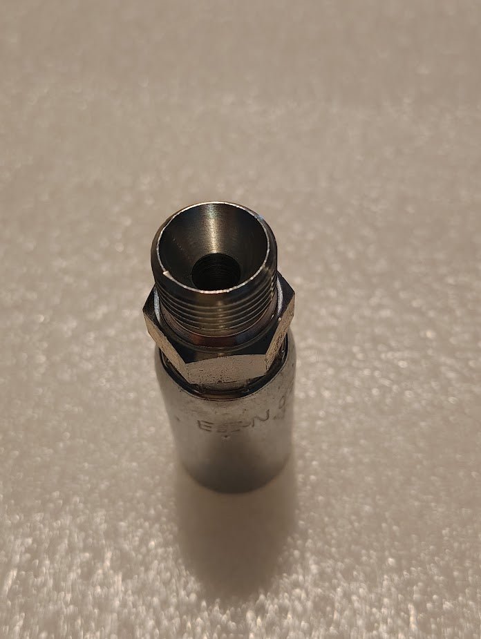 Close-up of a 06U-P56 Weatherhead 3/8 Hose x 3/8 BSPT Male Fitting standing upright on a textured surface. The fitting, with its hexagonal nut, reflects light from above and resembles premium fittings.