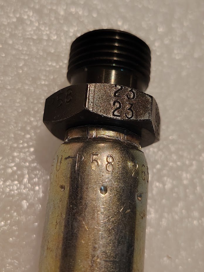 Close-up of the 06U-P58 Weatherhead fitting, featuring metallic threaded ends for 3/8 hose x 1/2 BSPT compatibility, with stamped numbers and markings. The background is a textured white surface.