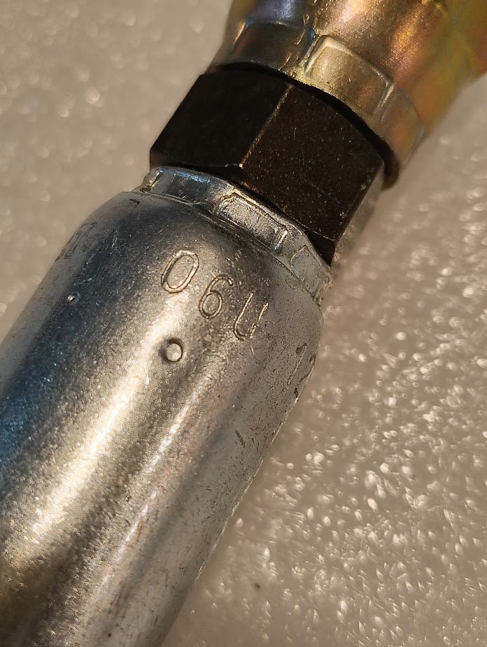 A close-up of a metal pipe with a hexagonal nut marked 06U-12C, similar to Parker fittings, against a textured, speckled surface.