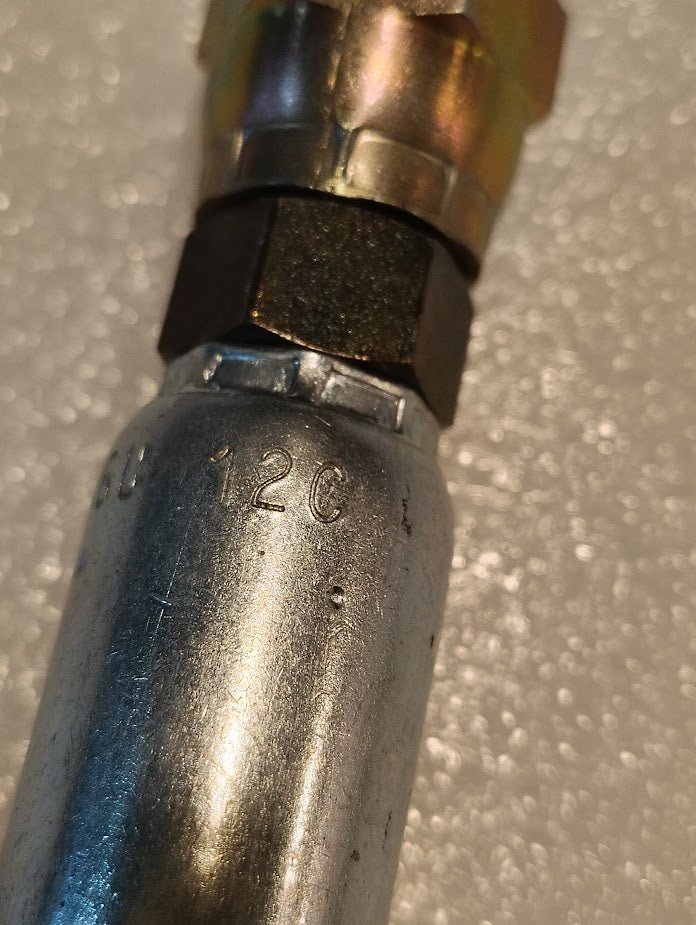 A close-up shows a metallic cylindrical object marked 12C L, featuring a top connected to a hexagonal nut and Parker fitting. The product, Weatherhead 06U-12C Hose x M18x1.5 DIN Female Fitting - Straight Swivel, rests on a textured, light-colored surface.