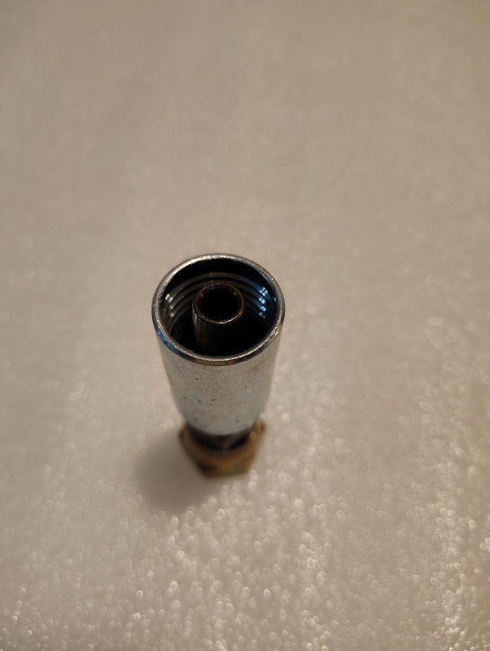 A close-up of a shiny Weatherhead 06U-12C fitting, resembling Parker fittings, stands upright on a textured white surface. Its metallic cylindrical form features a threaded interior with a silver finish and brass-colored base.