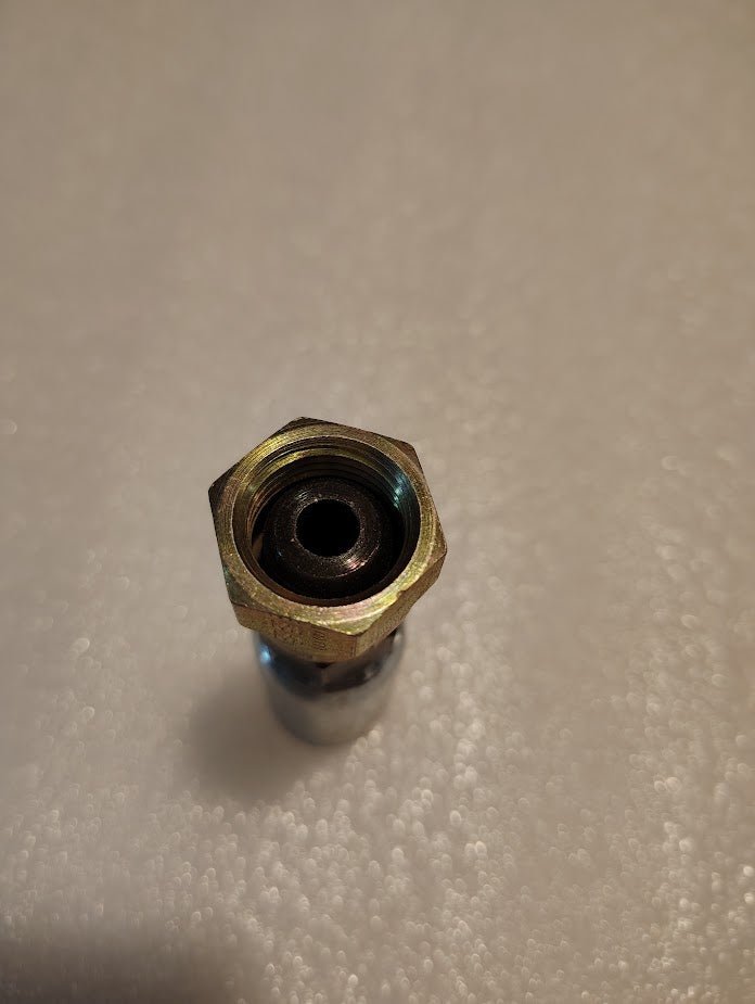 Close-up of a metallic Weatherhead 3/8 Hose x M18x1.5 DIN Female Fitting with a hexagonal nut top, similar to Parker fittings, placed upright on a textured white surface; the central hole suggests its part of a mechanical or plumbing component.