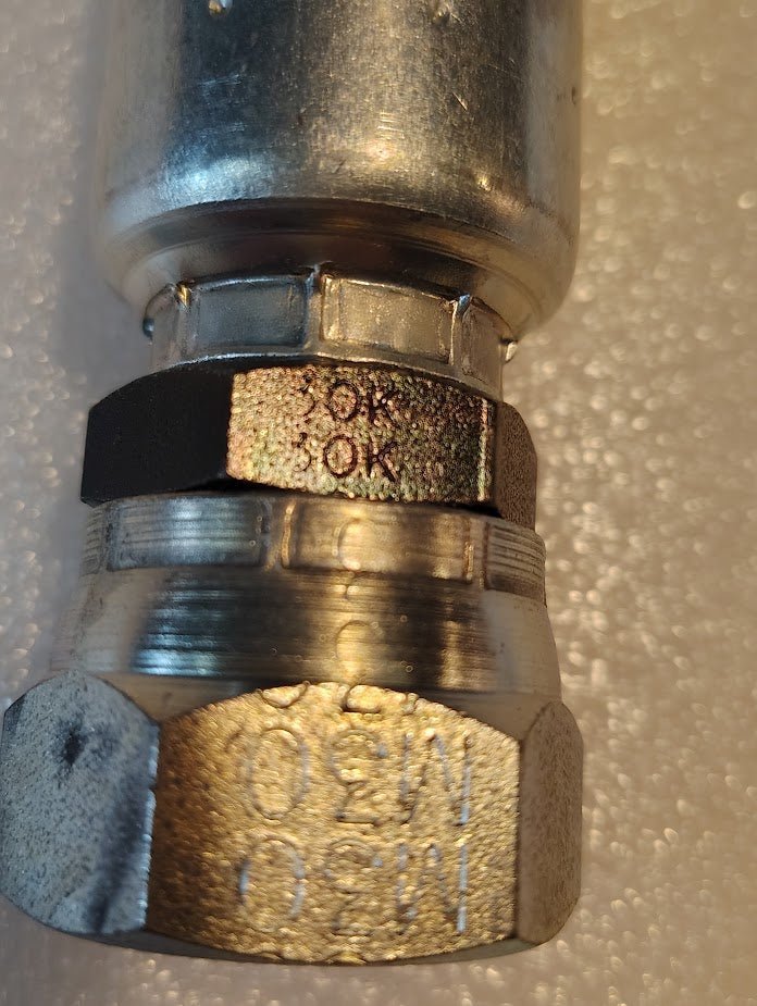 A close-up shows a shiny metallic connector engraved 30K and 250, resembling premium Weatherhead fittings. Compatible with 100R1 hydraulic hose fittings, it rests elegantly on a textured white surface. The Product Name is 12U-30K | Weatherhead 3/4 Hose x M30x1.5 Komatsu Female Fitting - Straight Swivel.