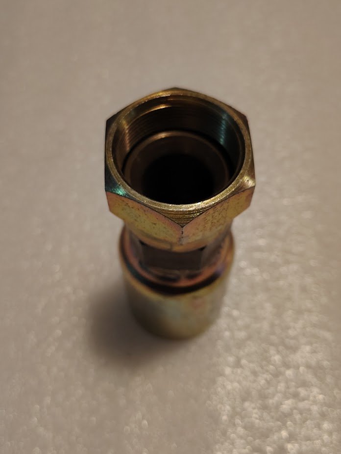Close-up of the Weatherhead 16U-33K fitting, a hexagonal, threaded 1 Hose x M33x1.5 Komatsu Female Swivel, against a textured white surface. The metal piece has a metallic finish with slight discoloration, highlighting its versatility with various fittings.