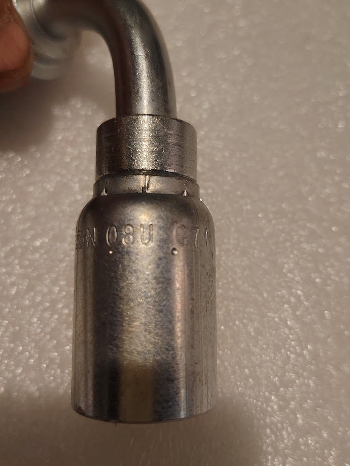 A metallic tool, similar to Weatherhead fittings, with a curved end rests on a textured white background. Engraved with numbers and letters, it resembles the 08U-G71 | Weatherhead 1/2 Hose x 1/2 Code 61 Flange Fitting - 90.